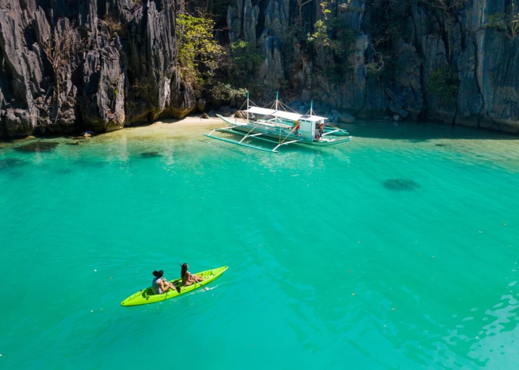 Man and woman kayak in green water under towering cliff with Filipino boat