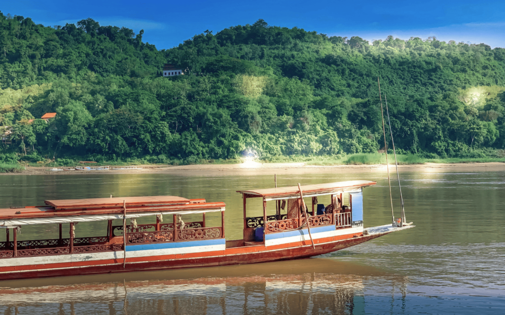 Traditional Laos long boat traveling brown river to Luang Prabang with green mountain in the background