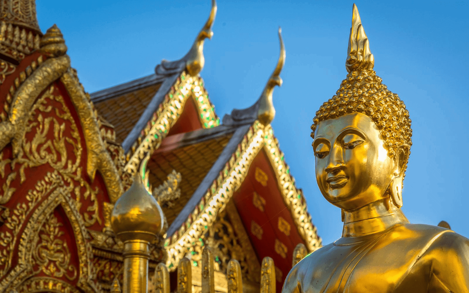 Gold and red Thai buddhis temple with golden buddha in front against a blue sky